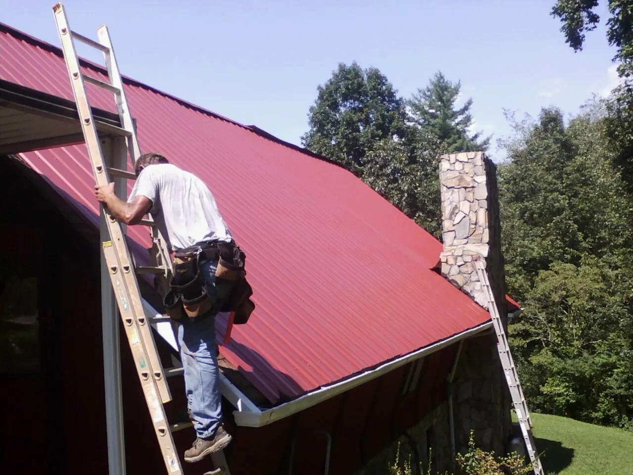 Roofer on a ladder beside a red metal roof and stone chimney — Soffit & Fascia Repair by Roofing Brothers in Leesburg